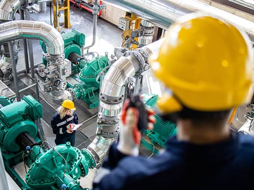 A man in a hard hat stands by a large industrial machine, observing a woman taking notes.