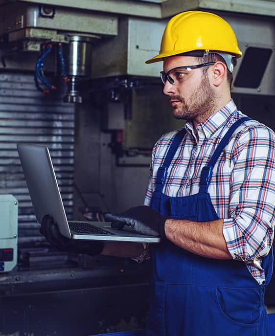 A construction worker wearing a hard hat and overalls operates a laptop.