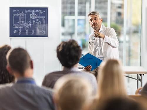 A man wearing a lab coat delivers a presentation to a group.
