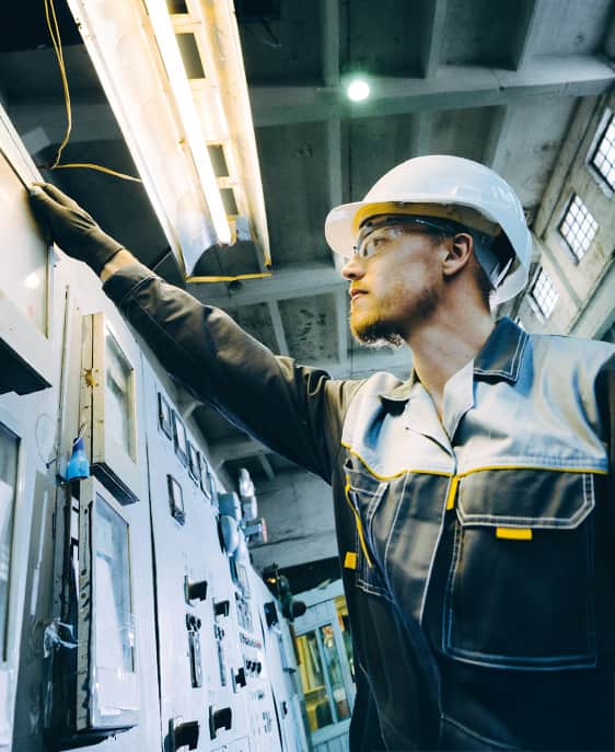 A man in a hard hat and overalls inspects a control panel.
