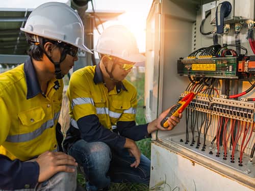 Two workers in yellow shirts and hard hats are collaborating on an electrical panel, ensuring proper installation.