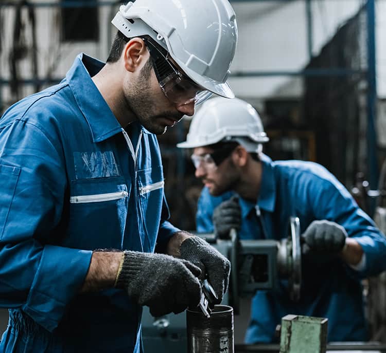 Two workers in blue coveralls and white hard hats operate machinery in a factory setting, focusing on their tasks with protective gloves and safety glasses.