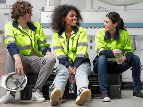Three individuals in safety vests seated on a bench, engaged in conversation outdoors.