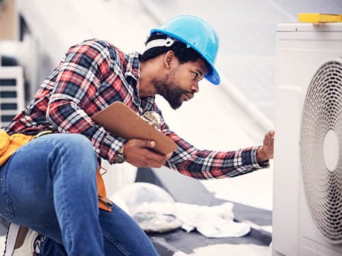 A man in a hard hat and blue shirt repairs an air conditioner.