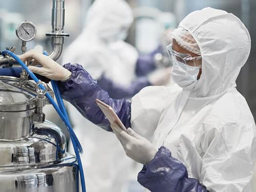 An individual wearing a white coat and protective equipment is engaged in machine maintenance in a lab.