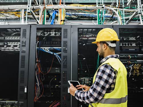 A man wearing a hard hat and safety vest stands in front of a server.