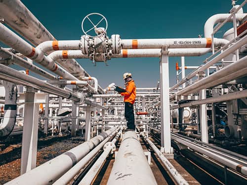 A man stands on a pipe at an oil refinery, surrounded by industrial equipment and structures.