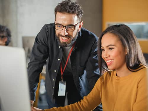 A man and woman smile while collaborating on a computer, showcasing teamwork and engagement in their work.