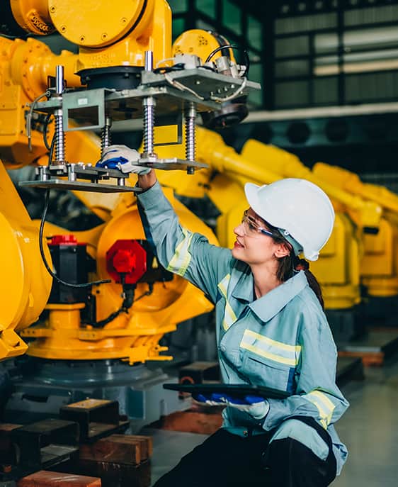 A woman in a hard hat and safety glasses is focused on working with a machine in an industrial environment.