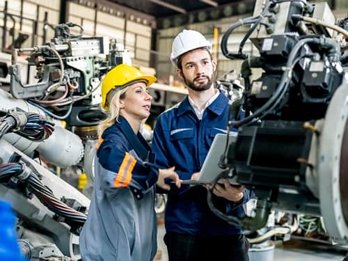Two workers wearing hard hats are positioned in front of industrial machinery.