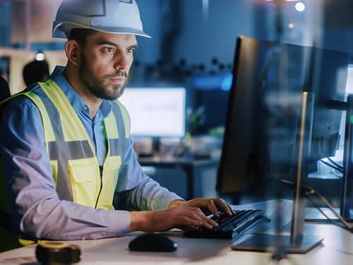 A man wearing a hard hat and safety vest is focused on working on a computer at a construction site.