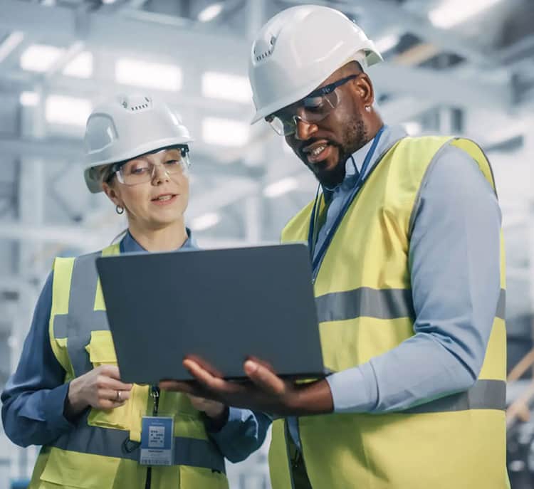 Two workers wearing hard hats and safety vests examining a laptop together.